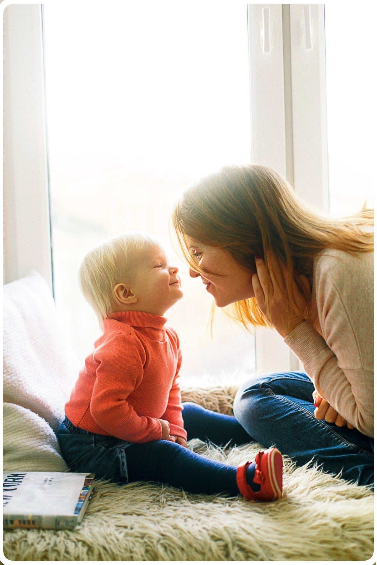 Woman and child looking at each other, noses touching. They sit in front of a window on a furry rug.