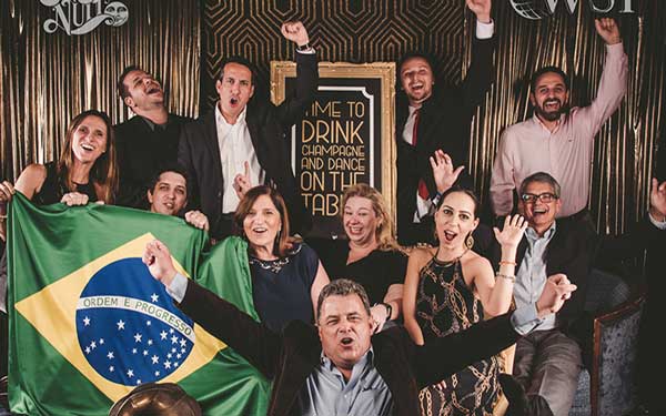 Group of people cheering at a bar, holding a Brazilian flag and raising their hands in celebration.
