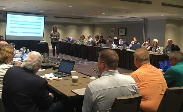 Conference panel meeting in a hotel room, with a presenter beside a projected slide and attendees seated at tables