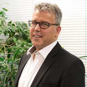 A man in a suit and glasses is standing in front of a window.