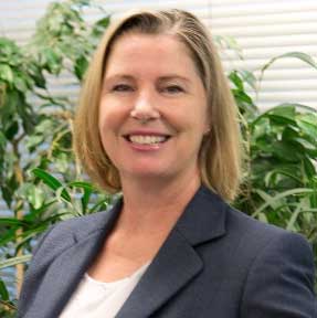 A woman in a suit and white shirt is smiling in front of a plant.