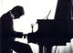 Silhouet van een pianist die een vleugel bespeelt; lessenaar zichtbaar. Zwart-witfoto.