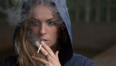 Person in a dark hooded jacket smoking a cigarette, with smoke drifting around their face.
