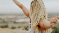 Blonde person with arms outstretched in a blurred outdoor landscape, facing away from the camera