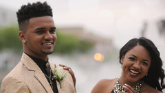 Two smiling people dressed for a formal outdoor event, with blurred lights in the background.