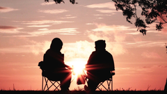Two people sitting in folding chairs at sunset, silhouetted against a bright orange sky.