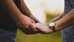 Two people holding hands, one wearing a wristwatch, outdoors with a blurred green background