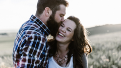 A smiling couple embracing outdoors in a sunlit field, with the man kissing the woman's forehead.