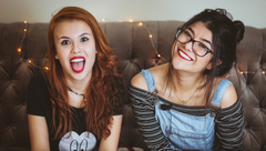 Two smiling friends sitting on a couch, one with red hair and one wearing glasses, in a cozy room with string lights