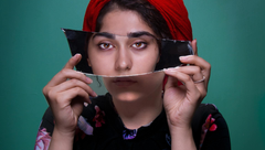 Woman in red headscarf holding a broken mirror, reflecting her face against a teal background