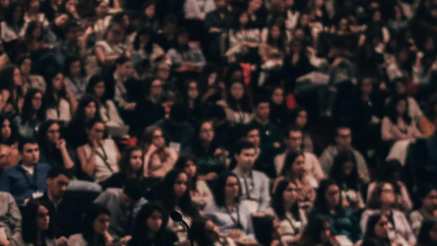 Crowded audience seated in a dimly lit venue, many people looking toward the stage