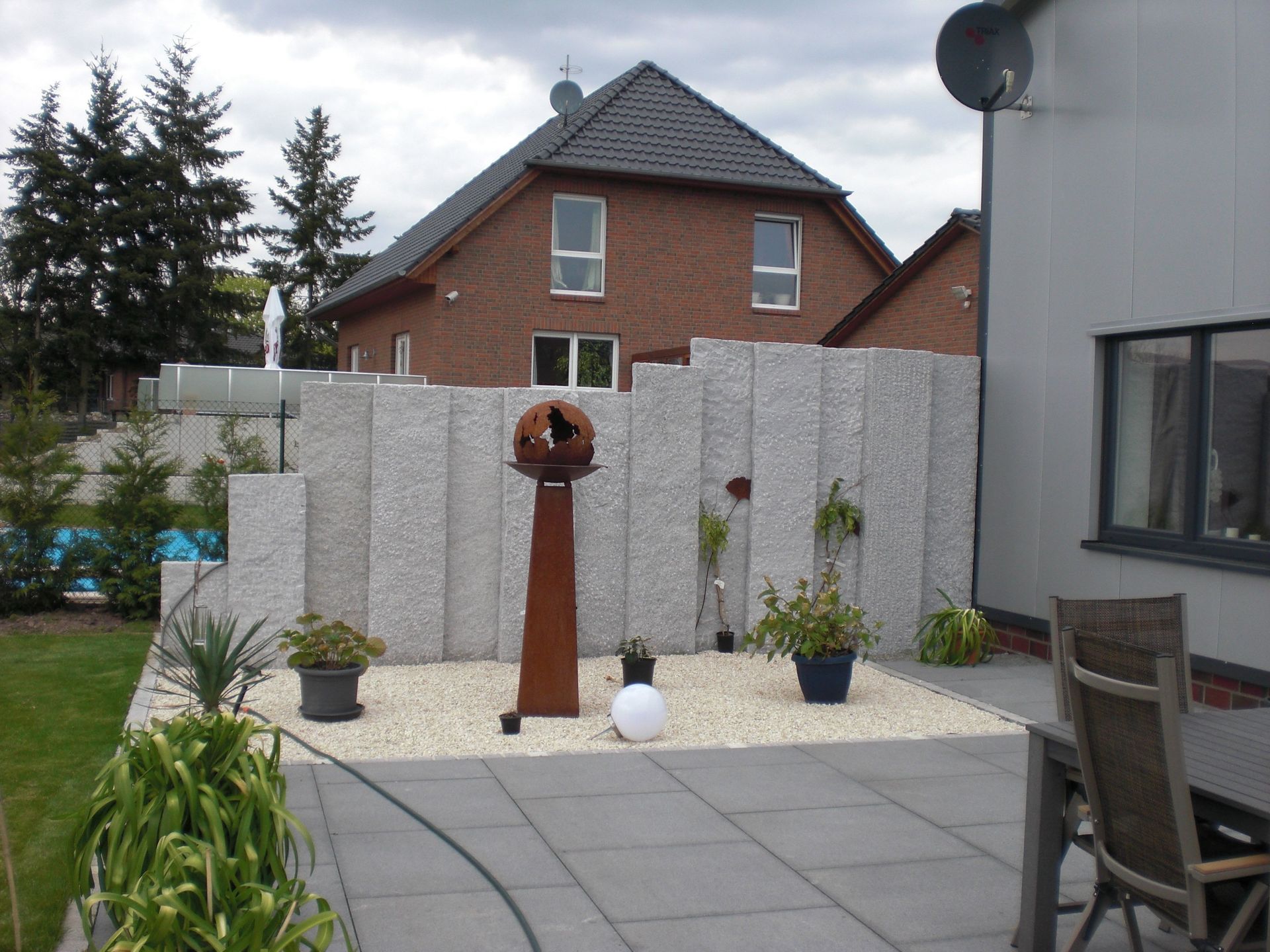 Terrasse mit hellen Platten, Granit-Sichtschutz und bepflanztem Gartenrand.