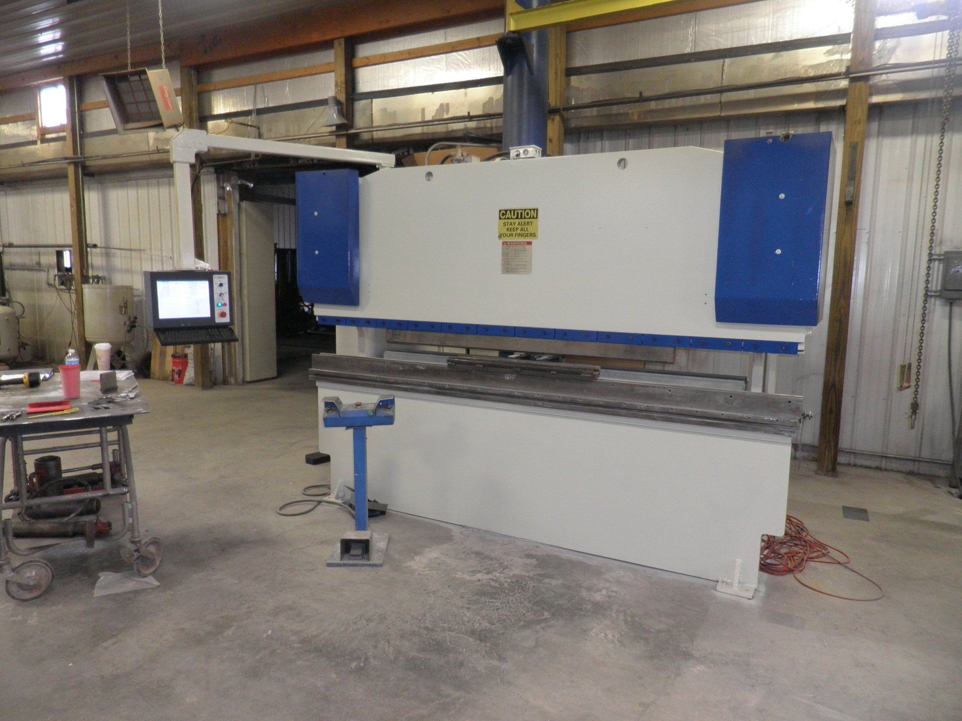Large industrial machine in a workshop, white with blue panels, beside a control screen and worktable.