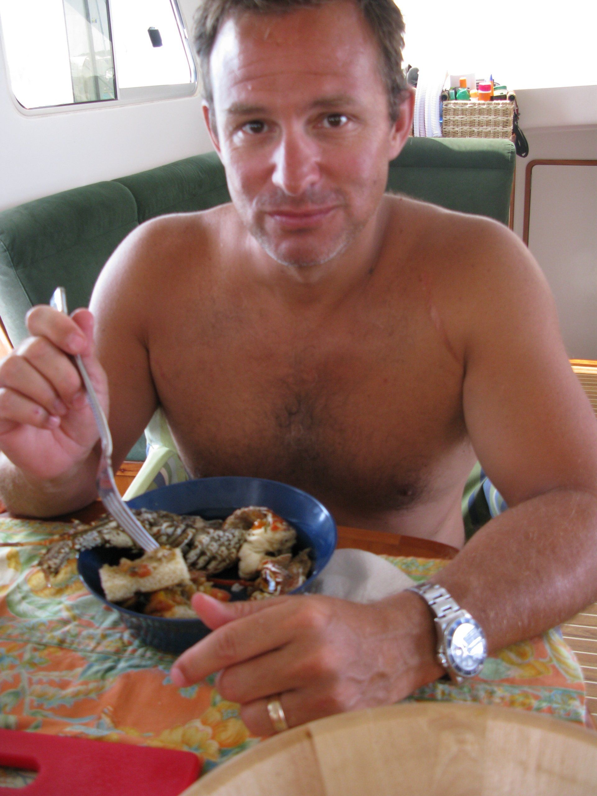 A shirtless man is sitting at a table eating a bowl of food