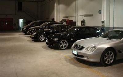 File di auto parcheggiate all'interno di un garage, illuminate da luci intense, tra cui berline nere e argento.