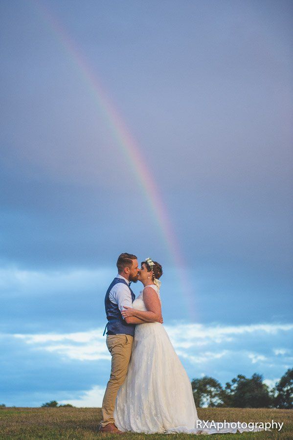 A bride and groom are kissing under a rainbow in a field.
