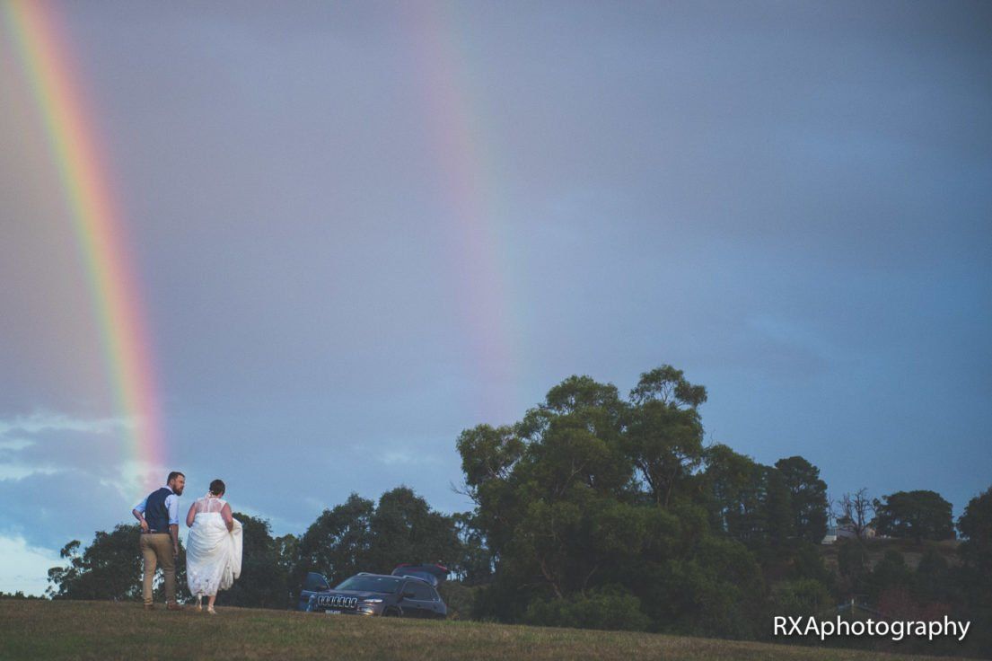 A bride and groom are standing under a rainbow in the sky.