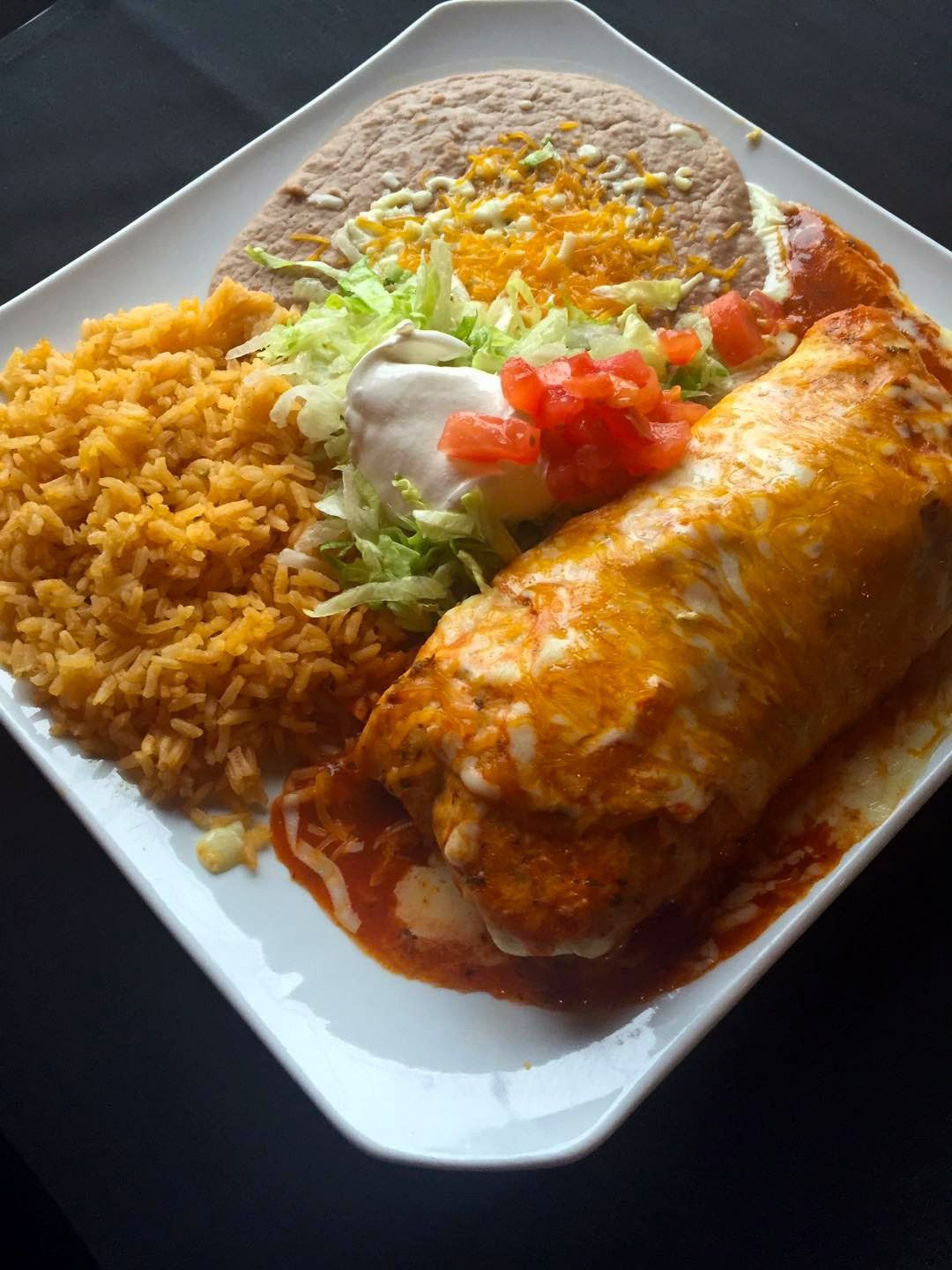 Plate of Mexican food: large burrito covered in sauce and cheese, with rice, beans, lettuce, tomatoes, and sour cream.