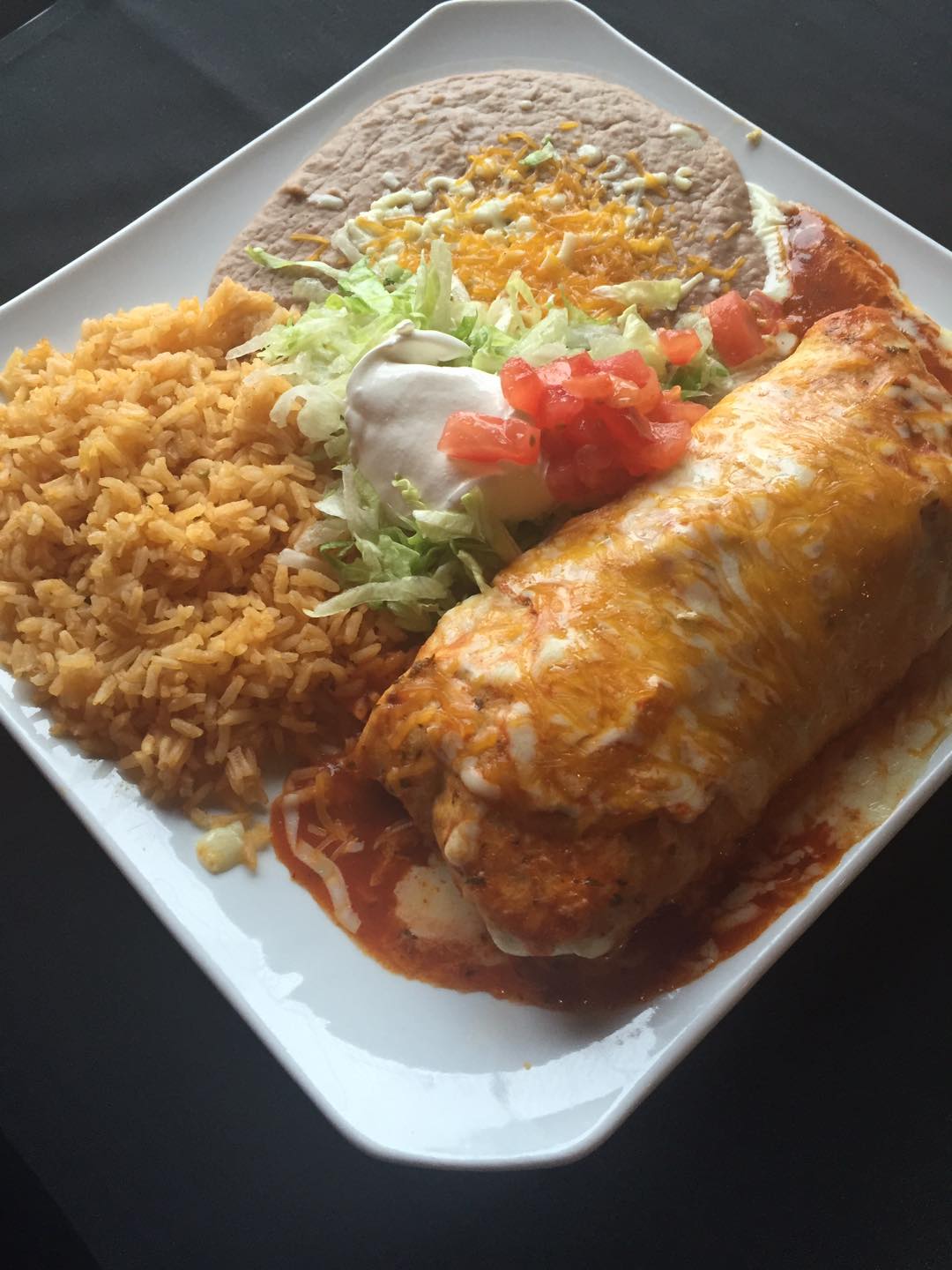 Burrito smothered in red sauce and cheese, with rice, beans, and toppings on a white plate.