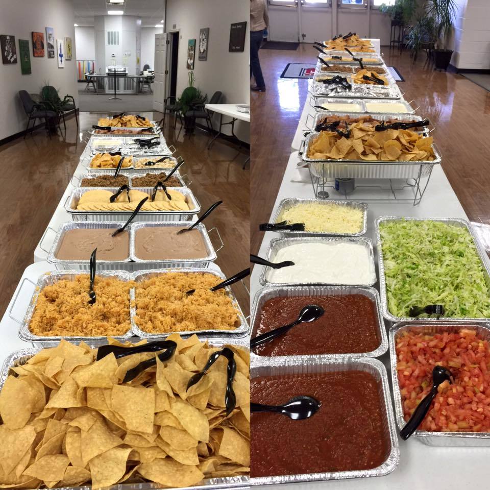 Buffet-style taco bar setup on long tables in a community hall. Chips, toppings, and fillings are in aluminum pans.