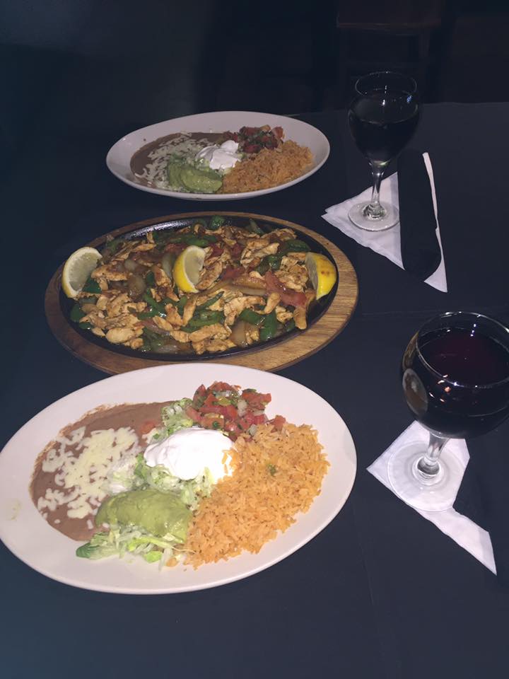 Table setting with two plates of Mexican food, a sizzling fajita platter, and two glasses of red wine.