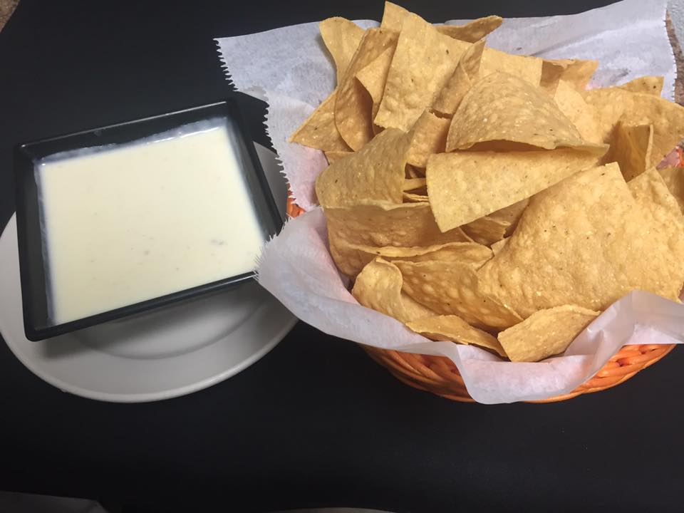 Chips and queso: basket of tortilla chips and a square bowl of white queso dip on a black table.