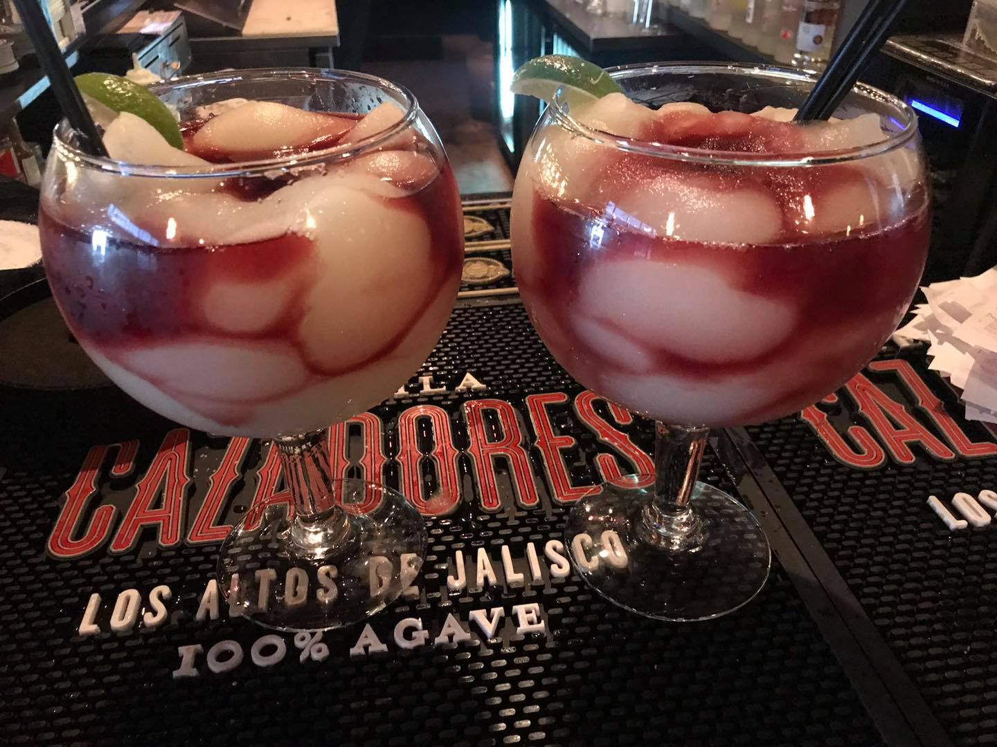Two cocktails in large globe glasses with fruit and a dark red swirl, on a bar.