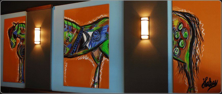 Paintings of colorful horses on orange backgrounds between warm wall sconces.