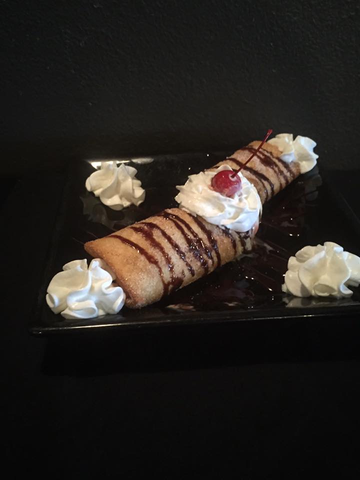 Fried dessert, cylindrical shape with chocolate drizzle, whipped cream, and cherry. Served on a black plate.