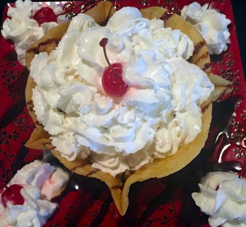 Dessert in a fried tortilla bowl, piled high with whipped cream and cherries, on a red plate.