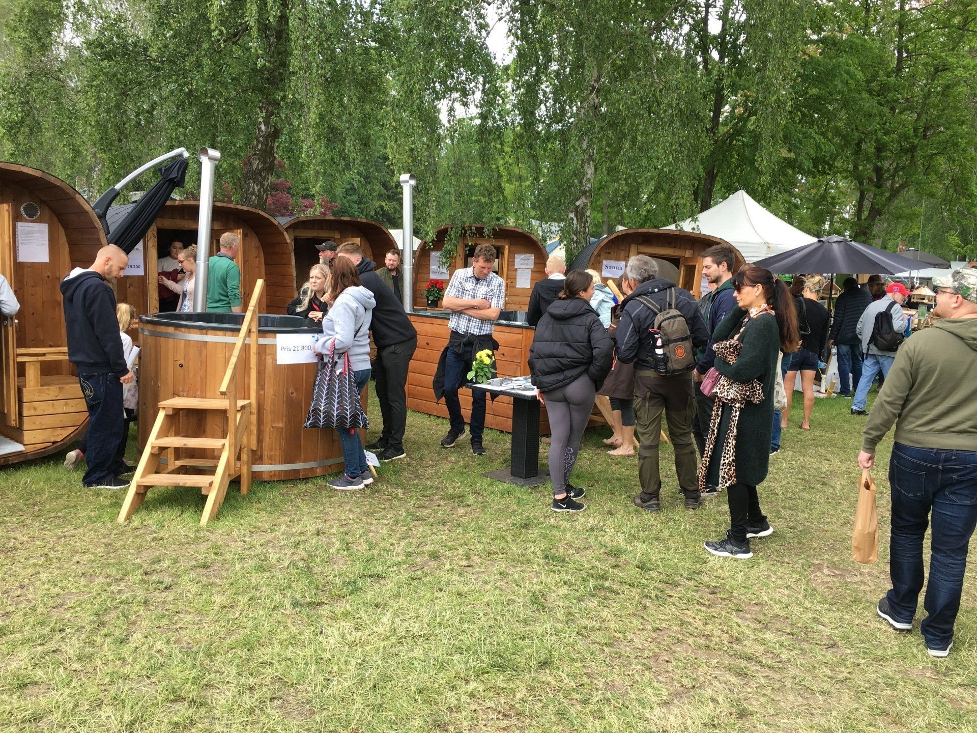 Folk samledes ved udendørs madboder af træ i en græsklædt park og snakkede under træer og telte.