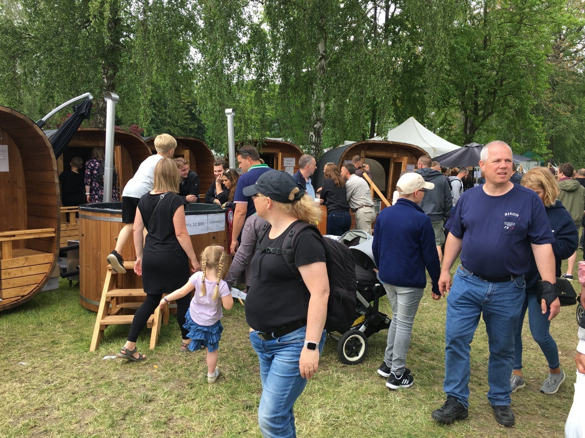 Folk samledes udendørs nær telte og en træstruktur på et græsklædt eventområde.
