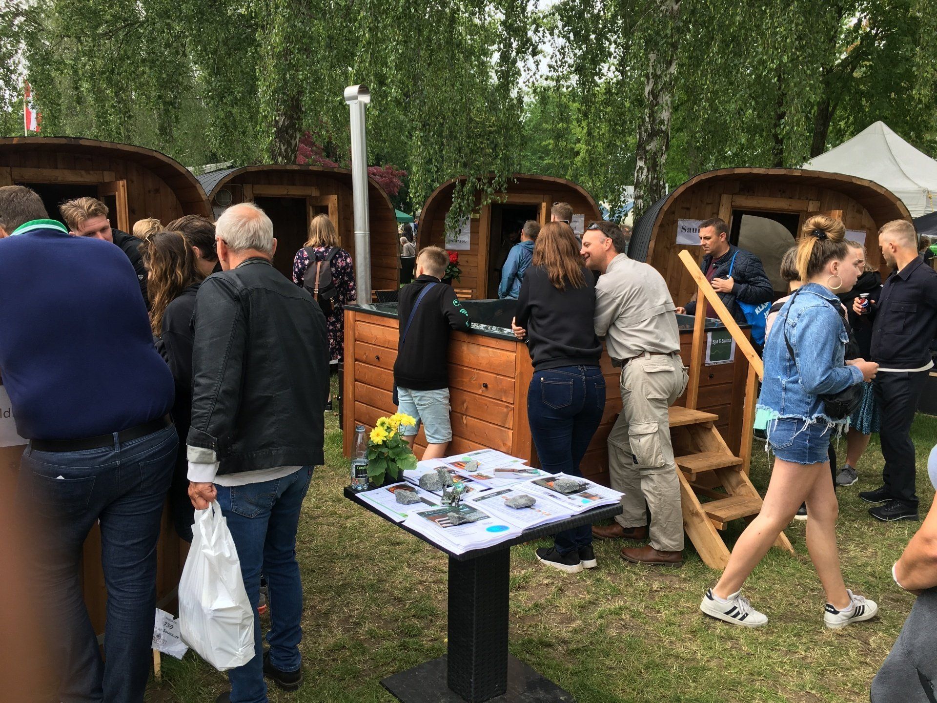 Folk kigger på udendørs markedsboder ved siden af ​​et bord med småting under træer.