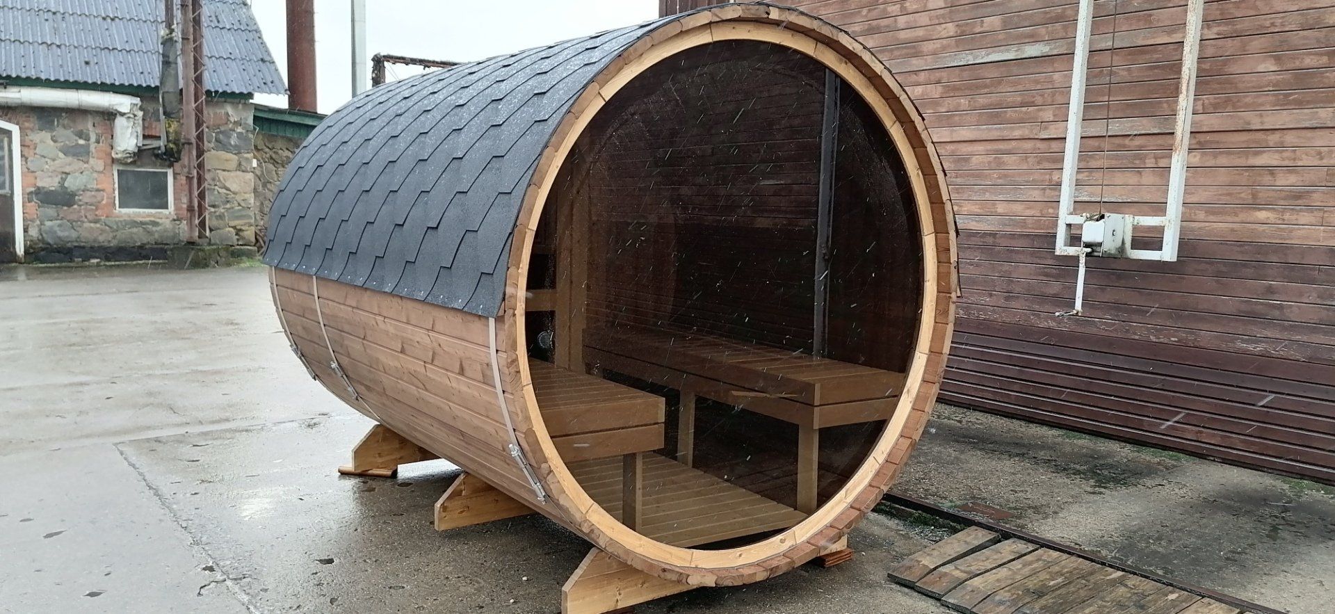 Tøndeformet sauna eller ly i træ på en våd fortov ved siden af ​​en murstensbygning