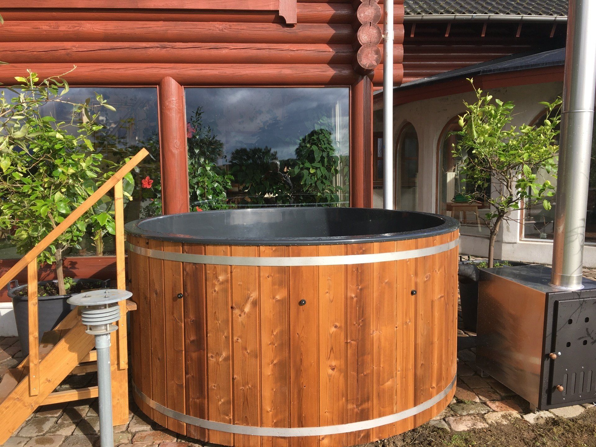 Rundt træjacuzzi på en terrasse ved siden af ​​en bjælkehytte med trapper og grønne områder i nærheden