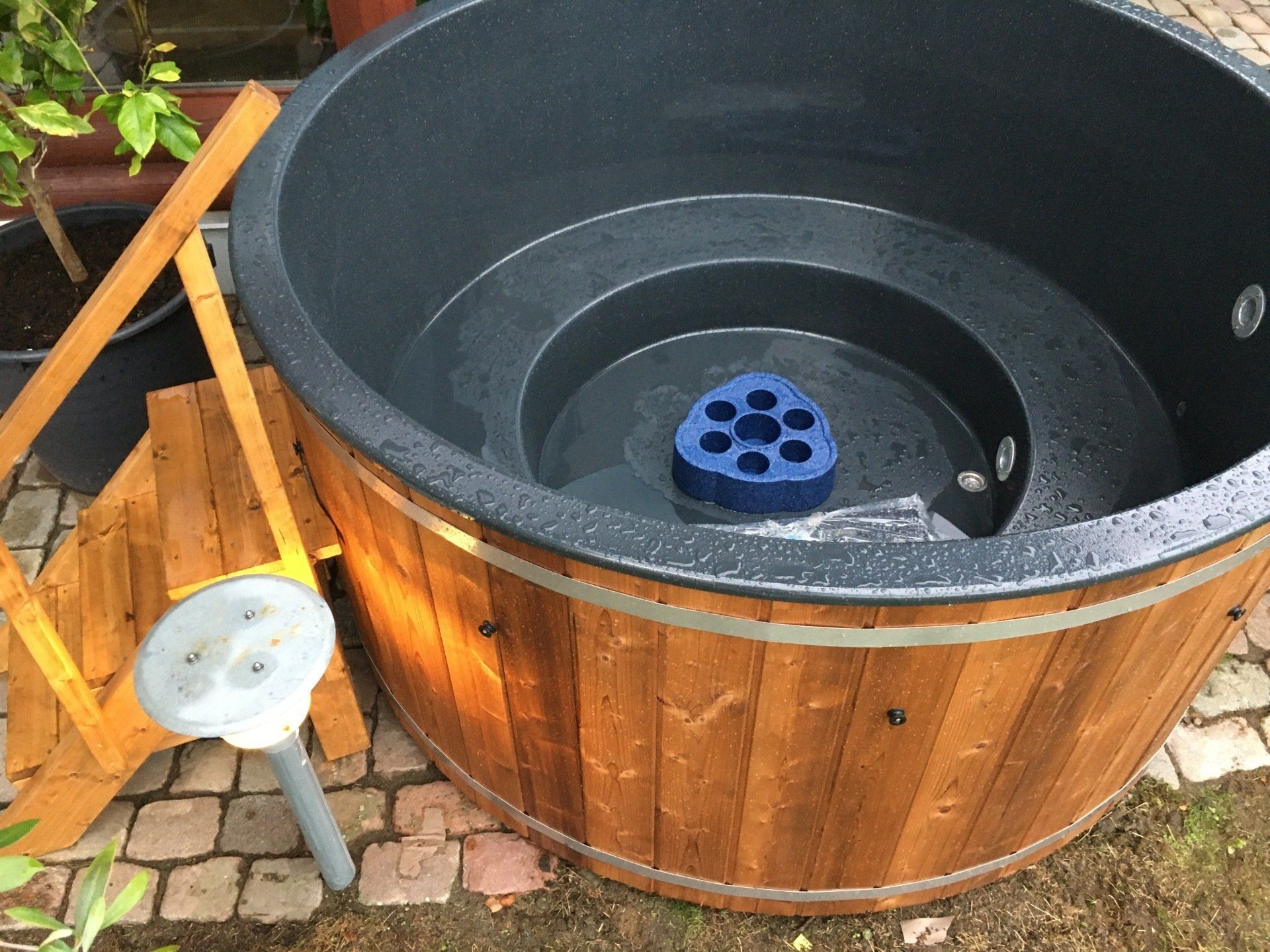 Træspabad med sort foring og blåt filter, udendørs på en terrasse ved siden af ​​en lille skammel.
