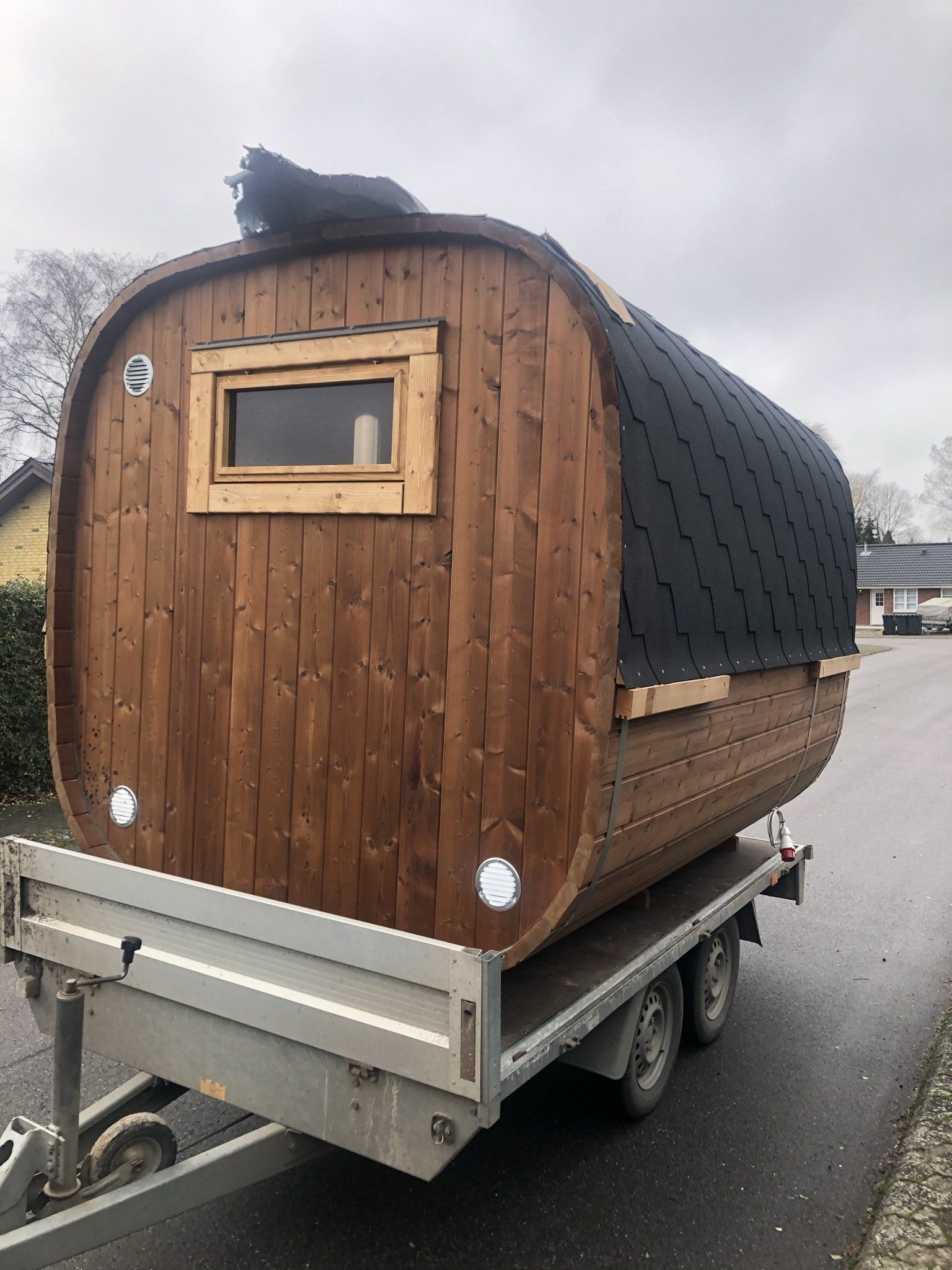 Træcampingvogn på en trailer med et lille vindue og et mørkt, buet tag, parkeret på en våd vej.