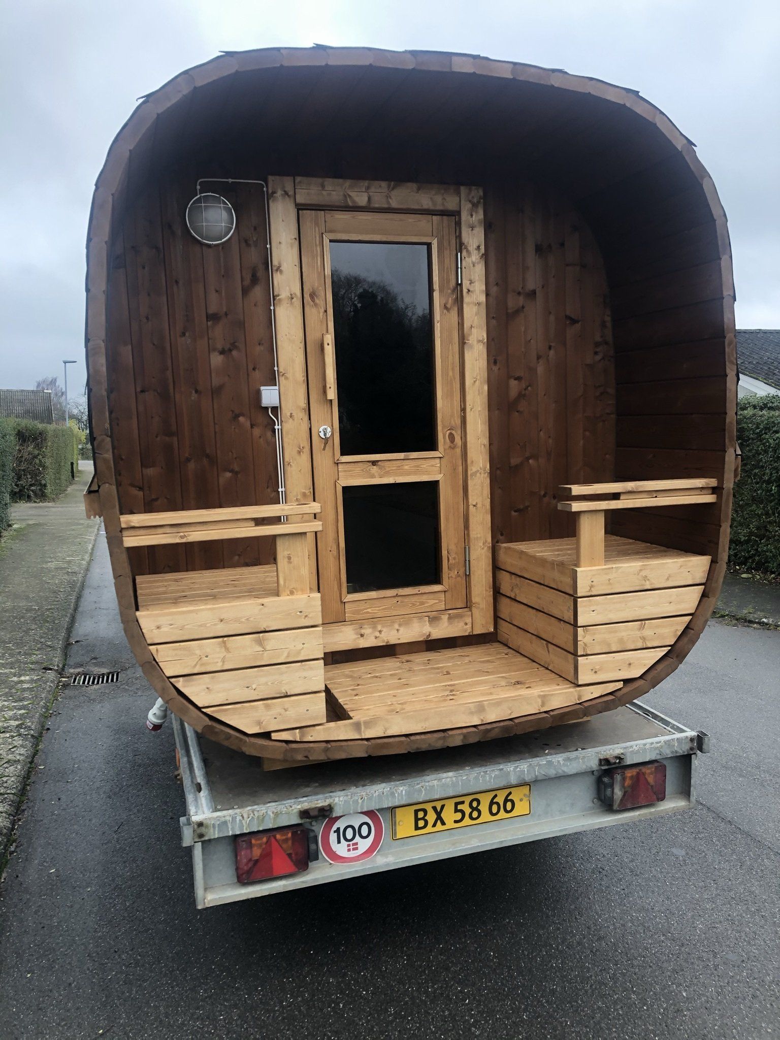 Lille træhytte på en trailer med et buet mørkt ydre og en hoveddør flankeret af små bænke.