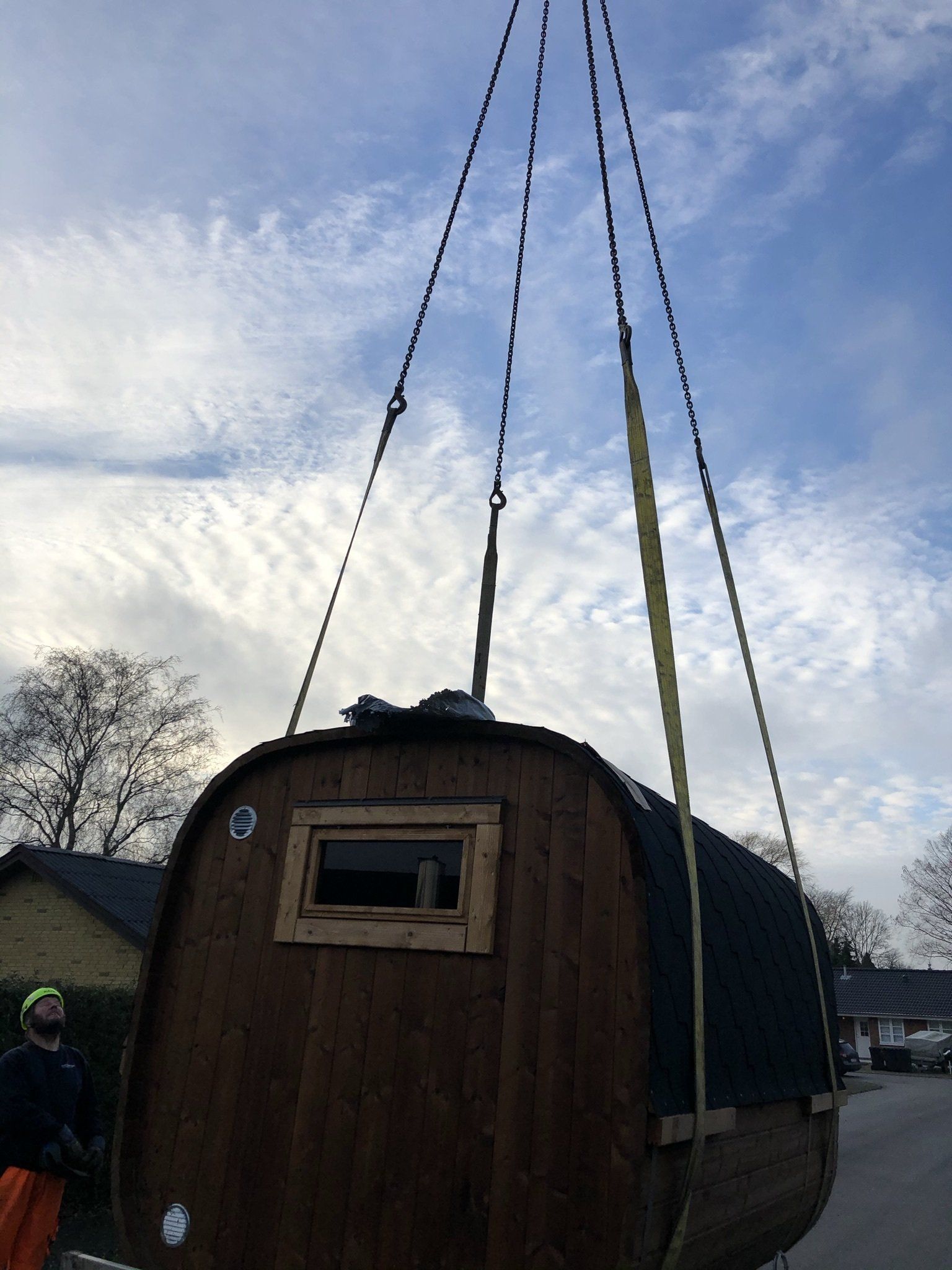 Lille træhytte løftes af krankabler mod en overskyet himmel