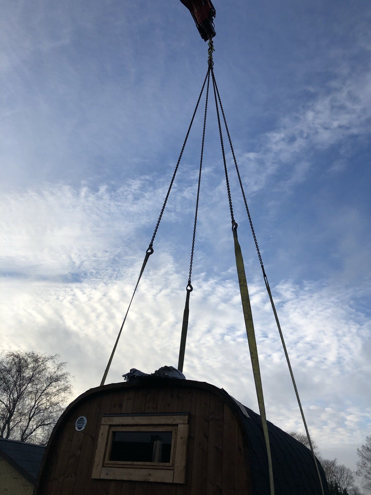 Lille træhytte løftet af kabler mod en blå himmel med skyer.