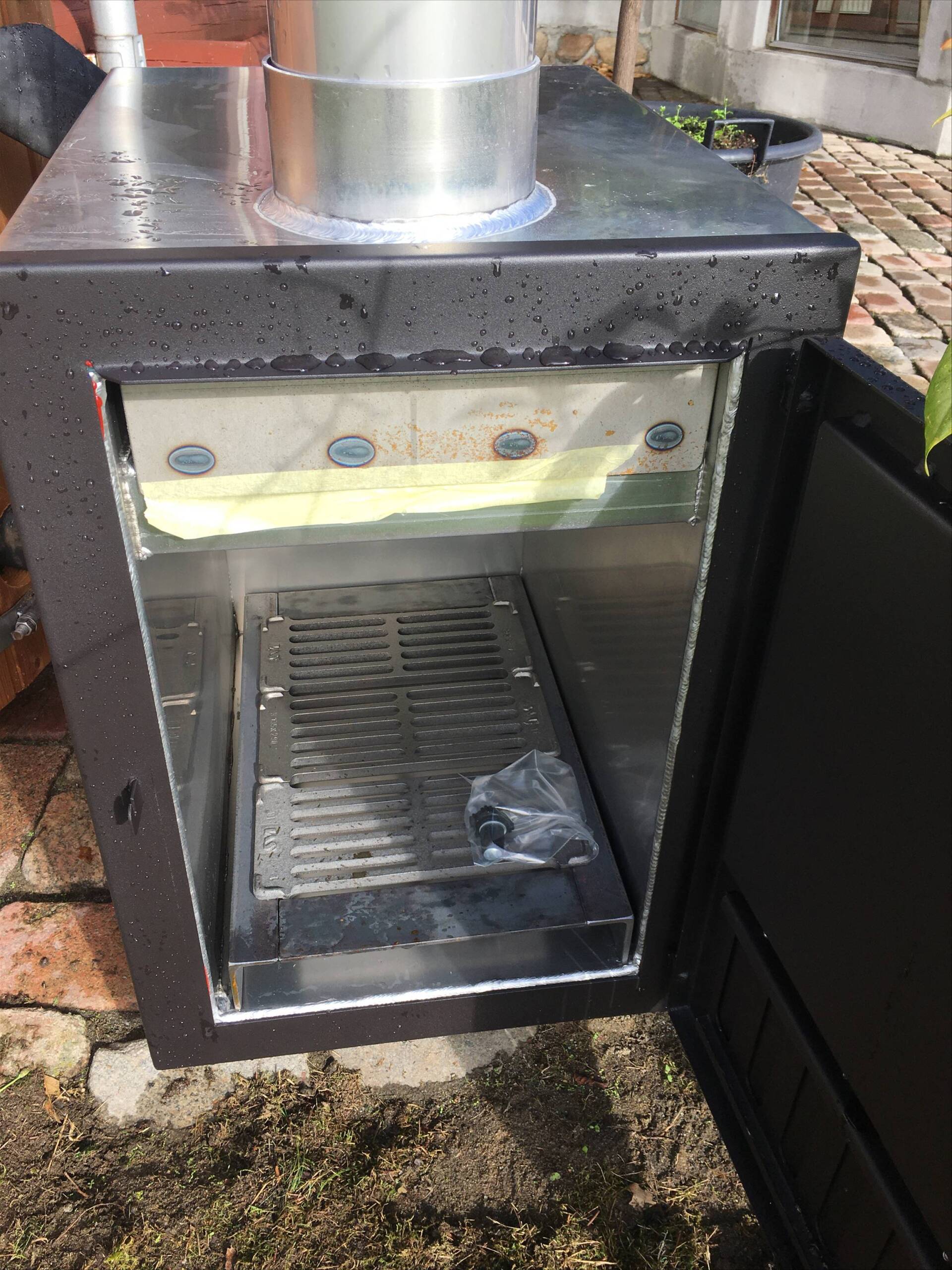Sort metal udendørs ryger eller grill med åben glaslåge og rustfri skorsten på en våd terrasse.