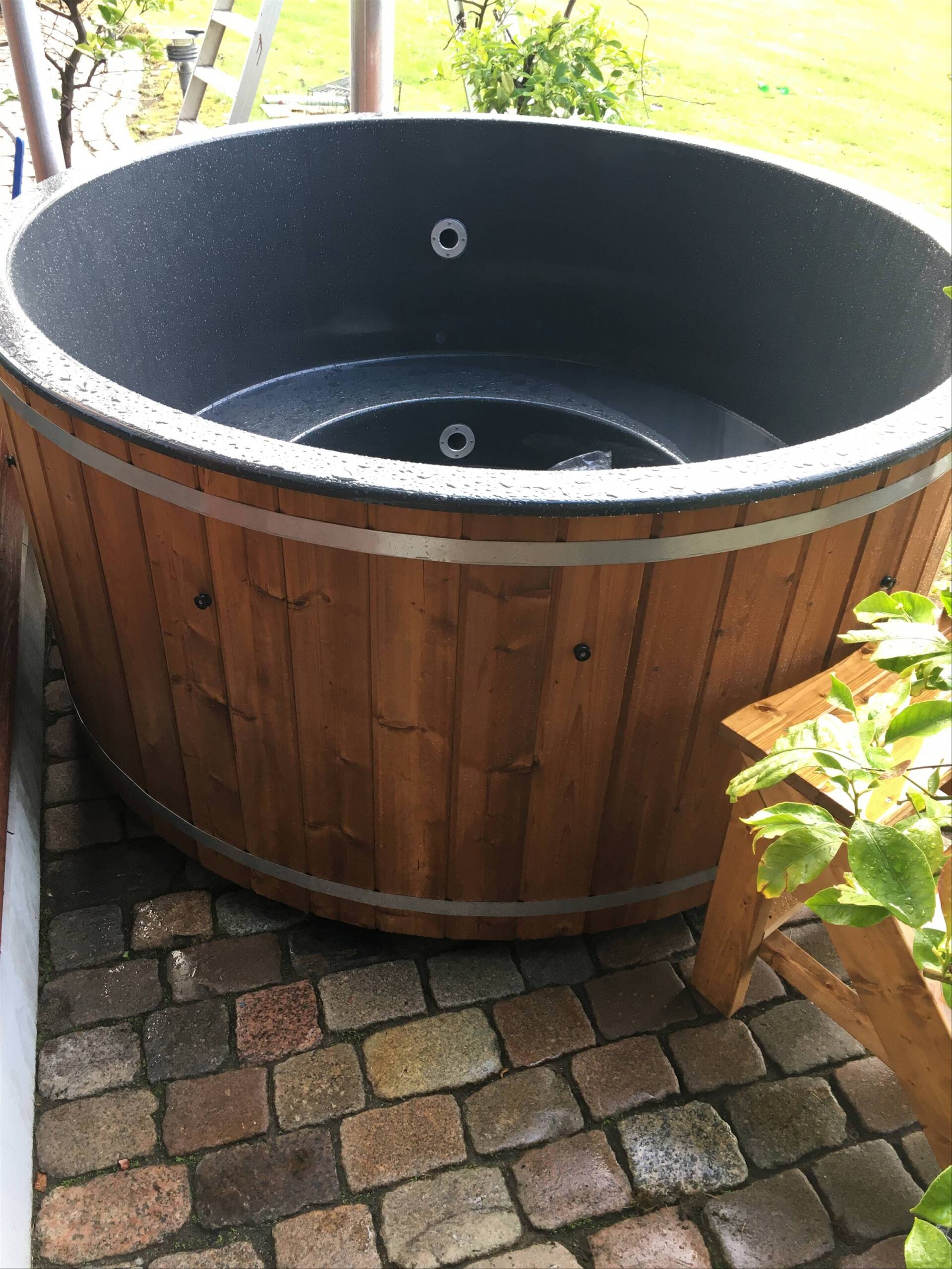 Rundt træjacuzzi på en murstensterrasse ved siden af ​​grønne områder, med sort inderside og metalarmatur.