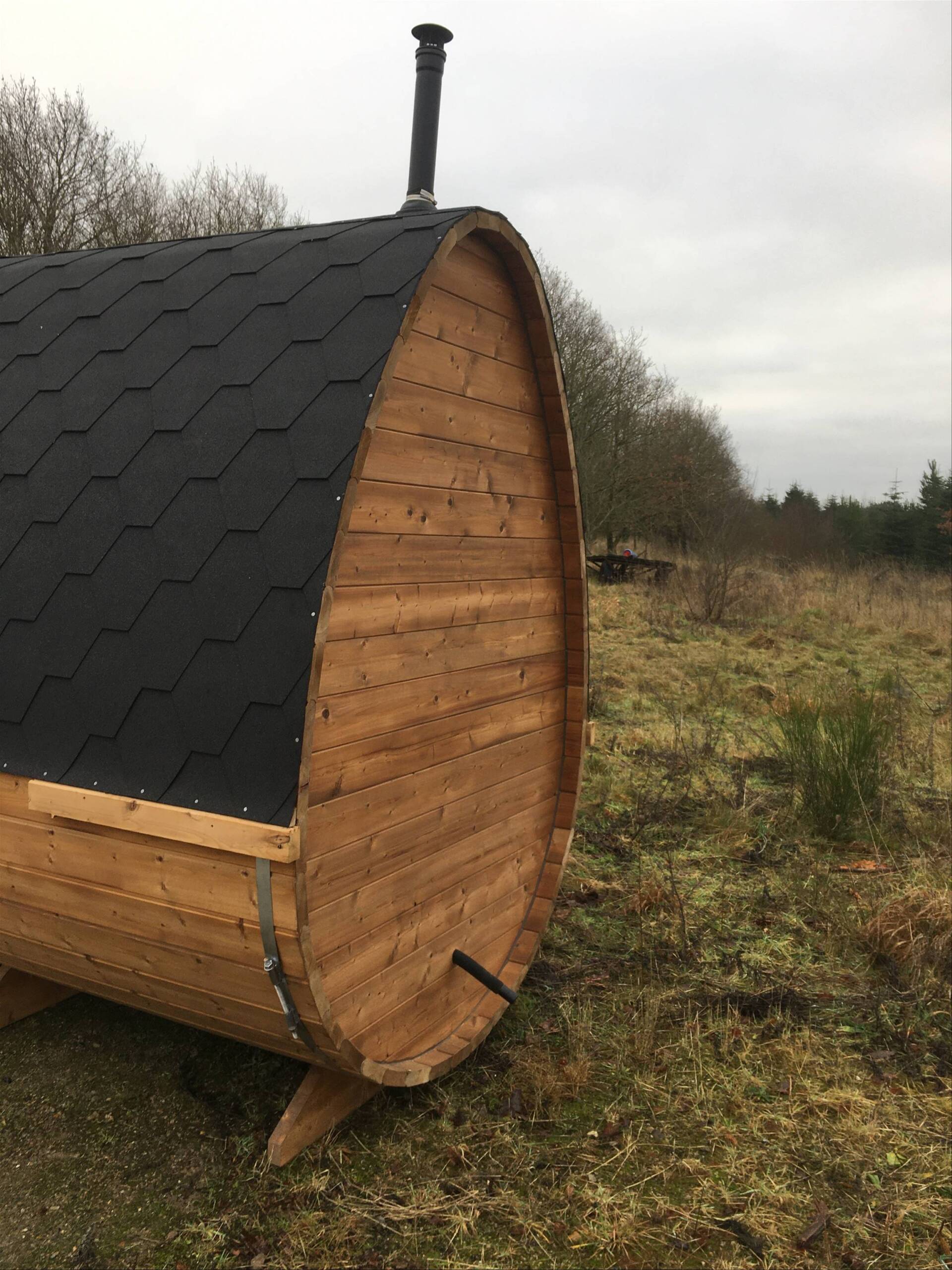 En lille trætønde-sauna med et sort tag af spånplader på en græsmark under en overskyet himmel
