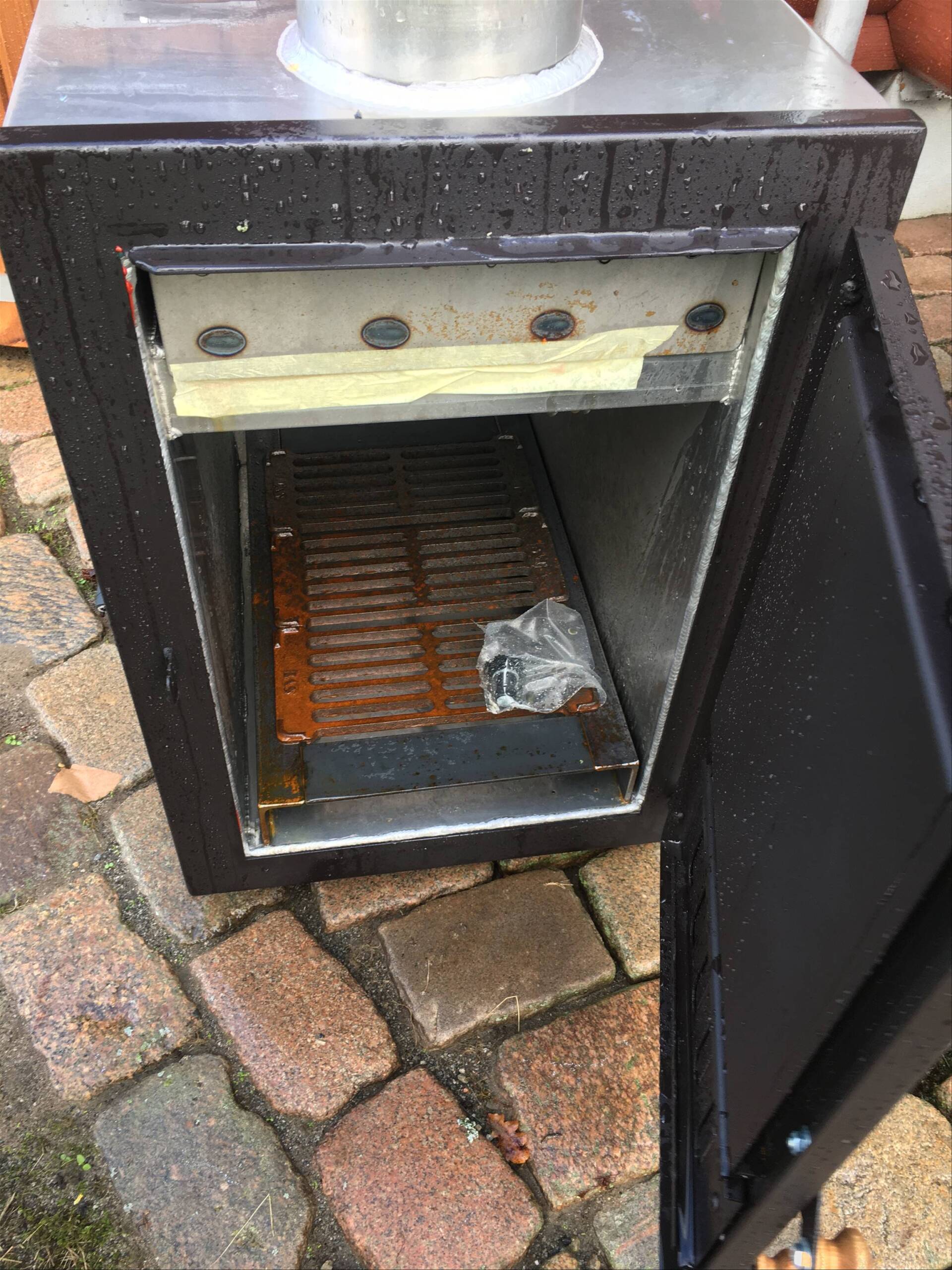 Åben metalovnslåge, der viser en rusten brændeovn på en murstensterrasse.