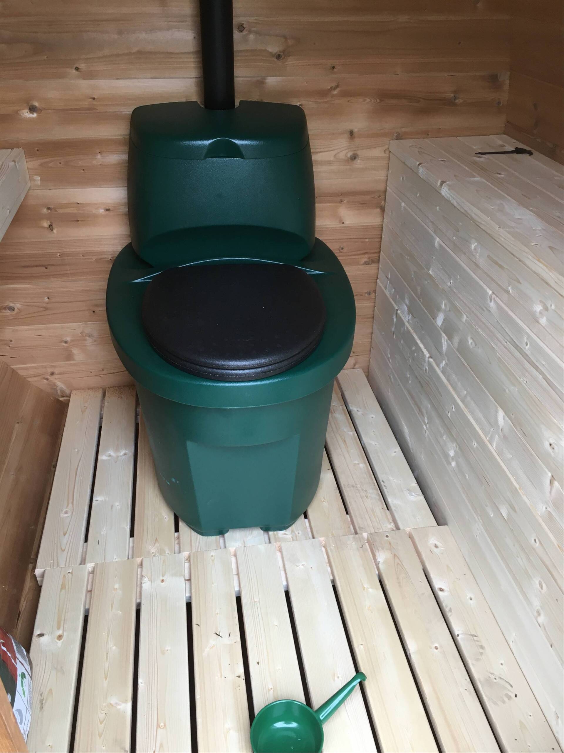 Grønt komposttoilet i et træskur med et sort sæde og en lille grøn skovl på gulvet.