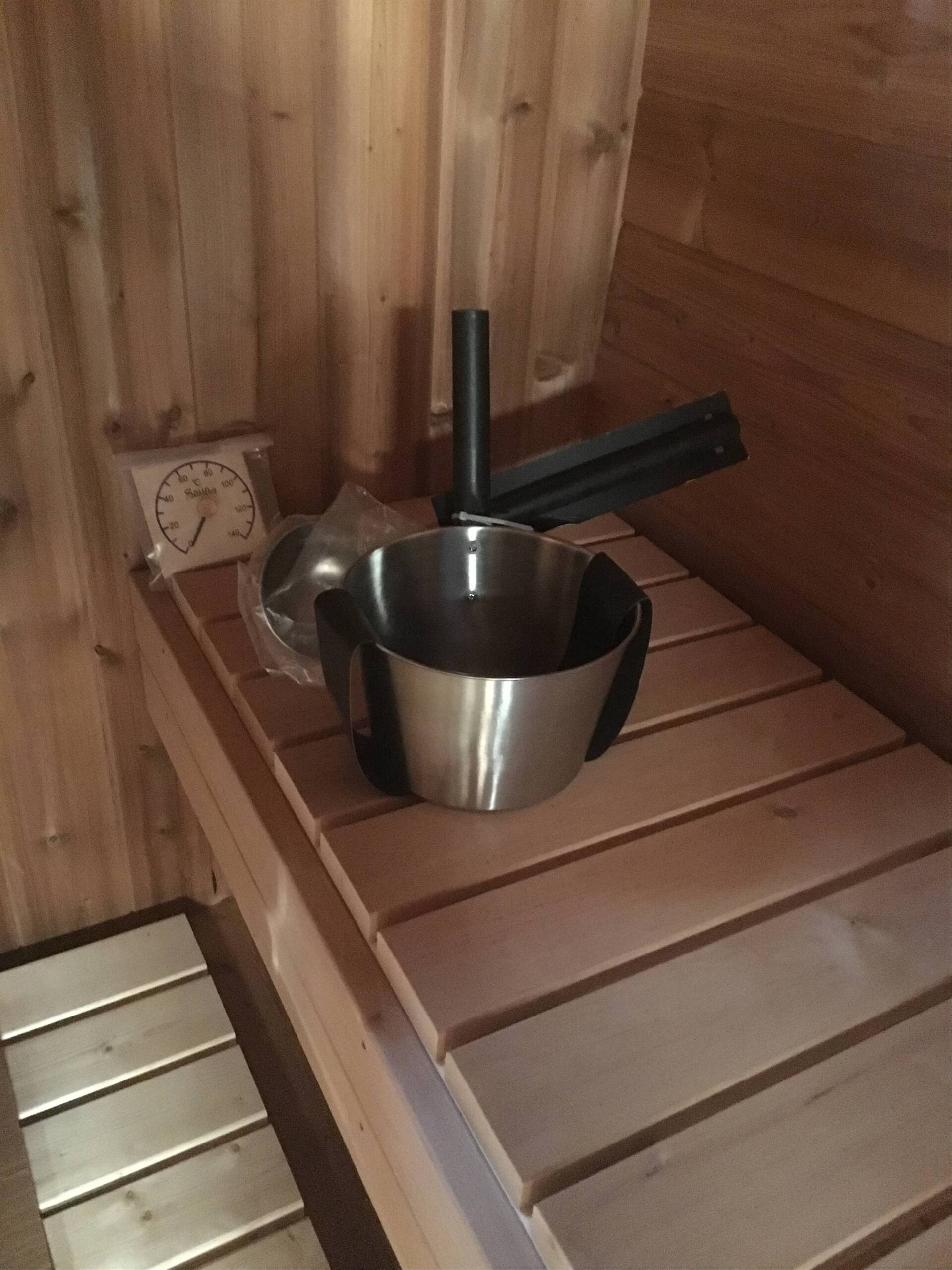 Metalspand med øse på en træbænk i sauna ved siden af ​​et termometer og komfurer.