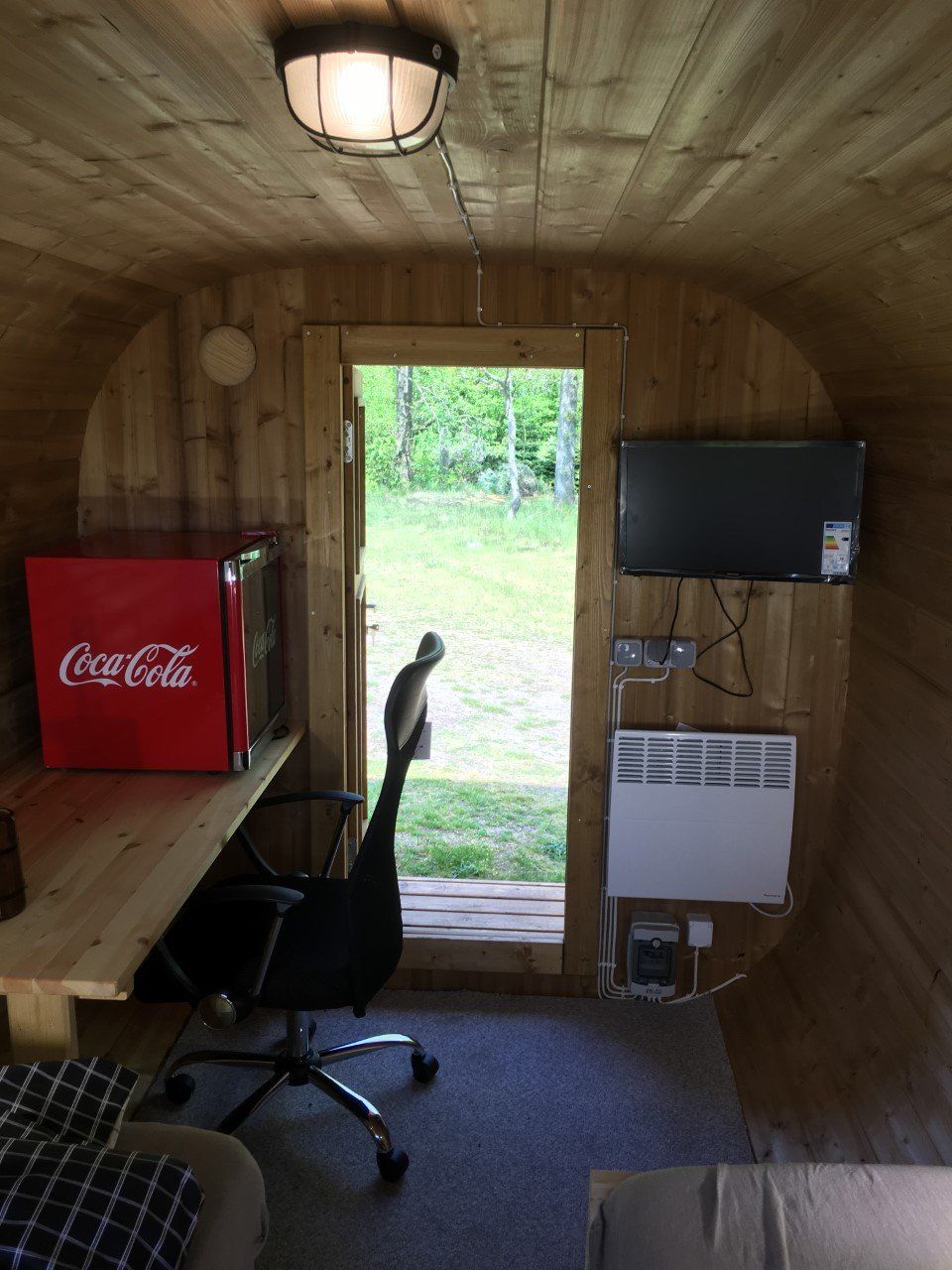 Lille træhytteinteriør med et skrivebord, Coca-Cola minikøleskab, væg-tv og en kat i den åbne døråbning.