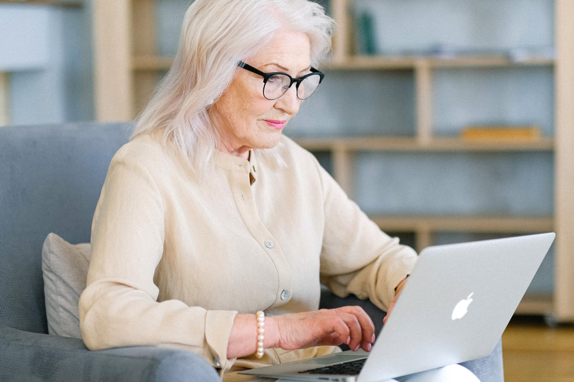 An elderly woman is sitting in a chair using a laptop computer.