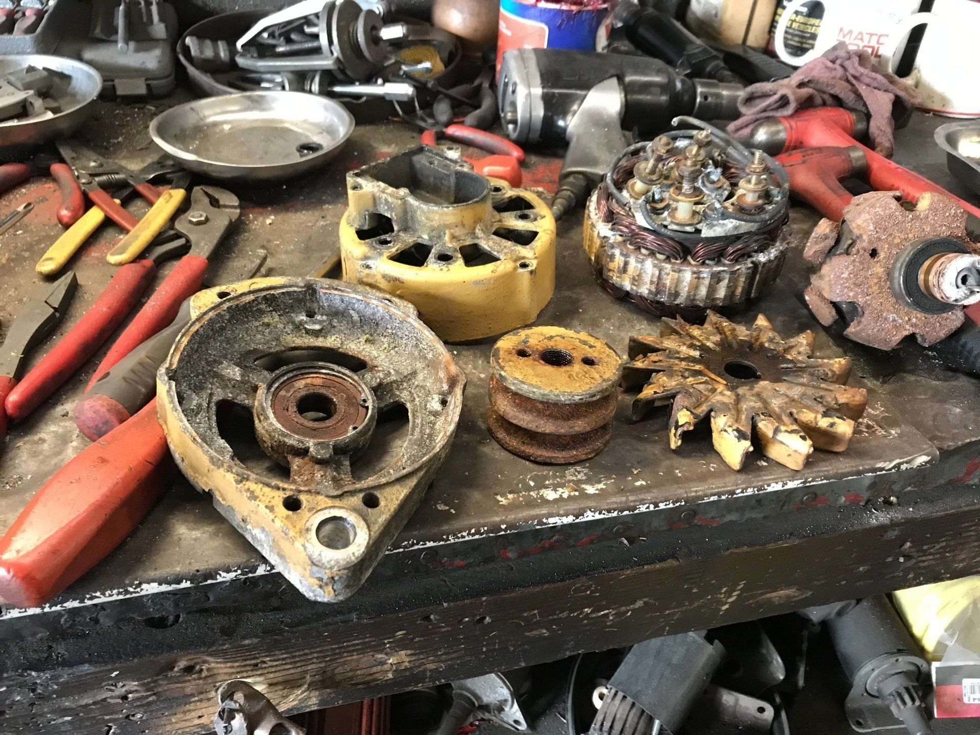 Scattered car alternator parts and tools on a cluttered workbench, with red-handled pliers and a rusty housing