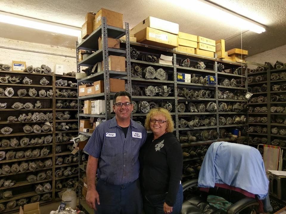 Two people standing in a storage room with shelves of organized metal parts and boxes.